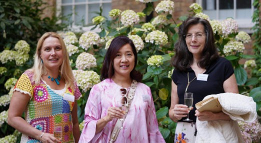 Three Goodenough College Alumni women with champagne glasses in front of flowers